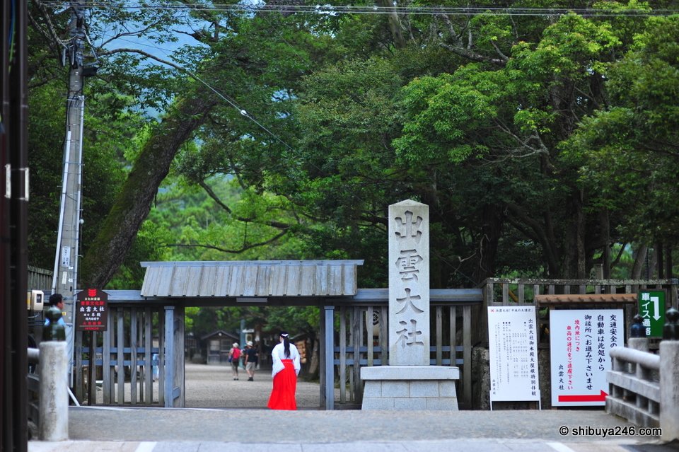 A shrine gate with a stone pillar and a shrine maiden in red walking past