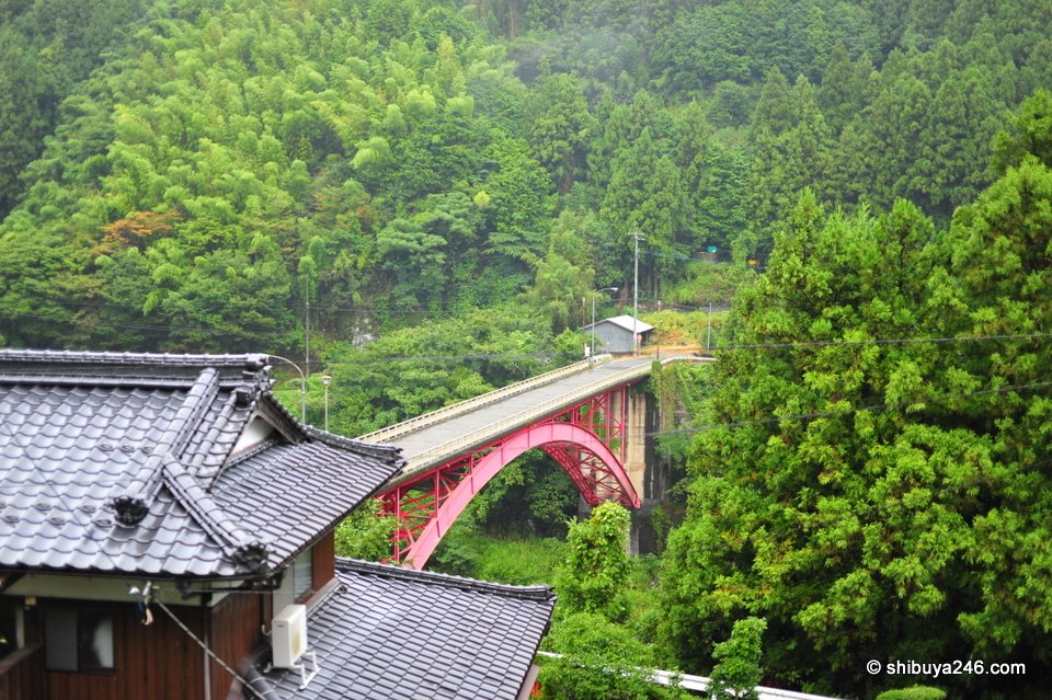 A red arch bridge across a green valley behind tiled roofs