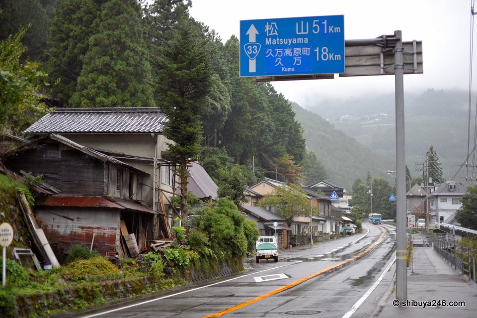 An old house beside the road with a blue sign to Matsuyama