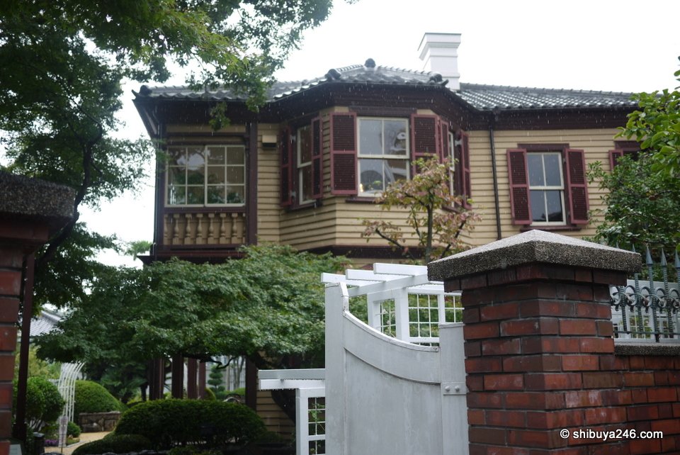 A dark wooden house with a balcony behind a brick gate