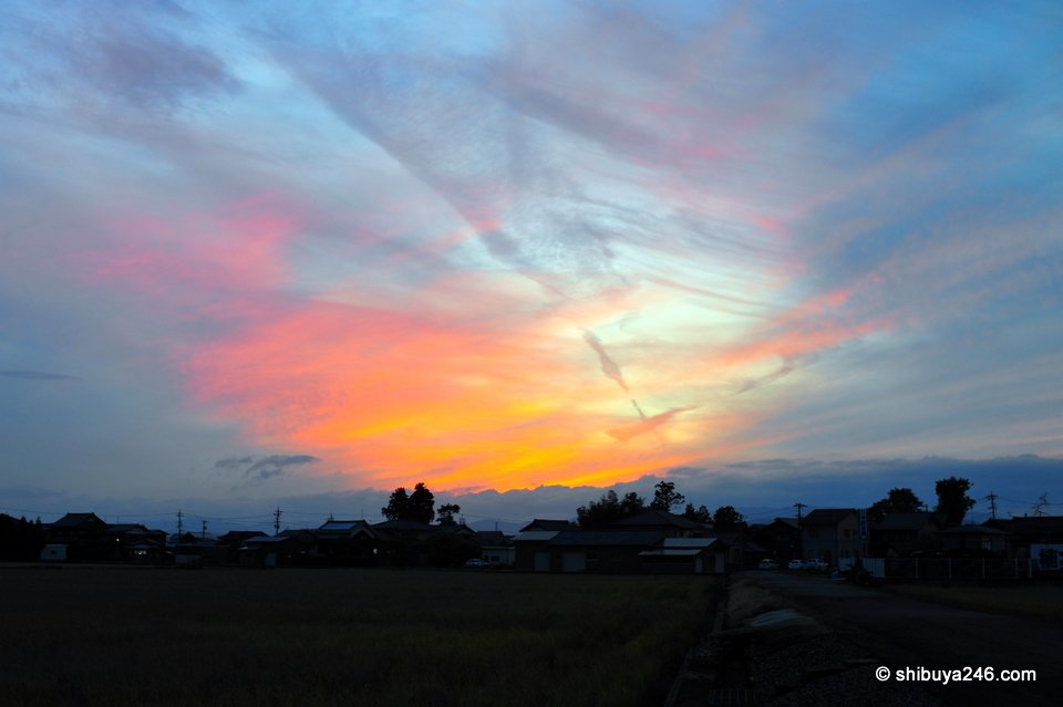 A wide sky of blue and orange over flat fields and houses