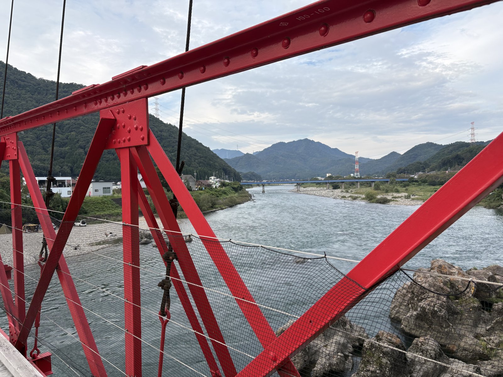 The red steel frame of a suspension bridge with the river and mountains beyond