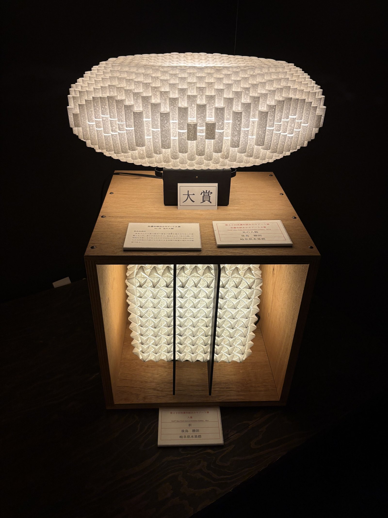 A ring-shaped lamp of folded paper on a wooden stand with a grand prize sign