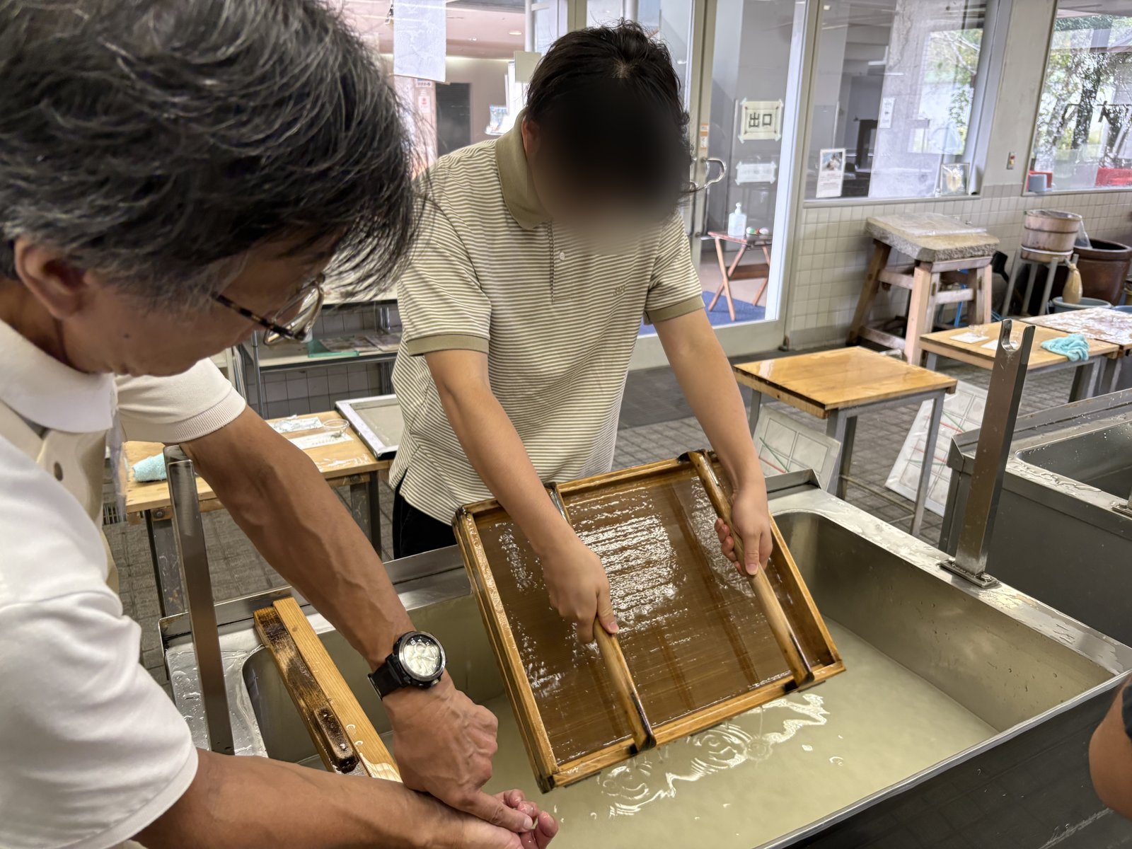 A young man tilting a wooden frame of wet paper pulp over a vat, with a staff member helping